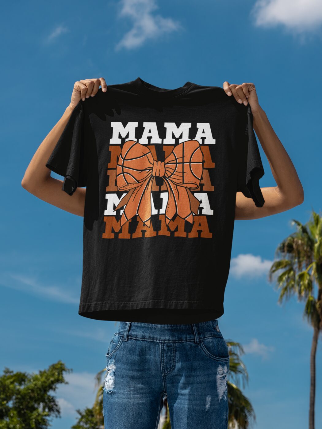 Someone holding up a black t-shirt printed with the word MAMA in orange basketball lettering, against a bright sky and palm trees.
