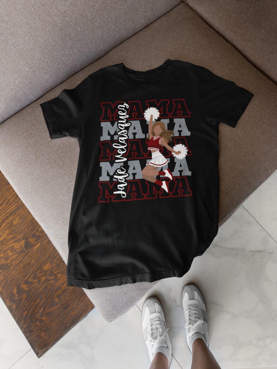 A black t-shirt laid out on a wooden floor, printed with a repeated MAMA design in red and white cheer lettering.